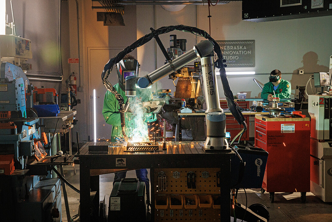 A Nebraska Innovation Studio member watches the welding robot while checking the programming on a tablet.