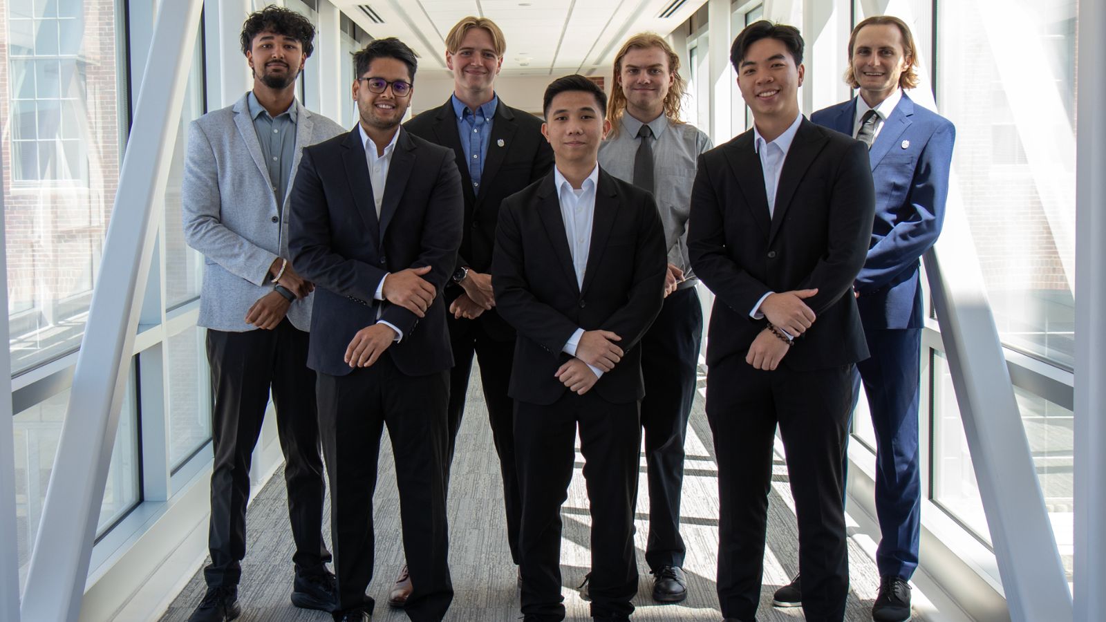 Seven men wearing formal suits stand confidently in an indoor hallway with glass windows.