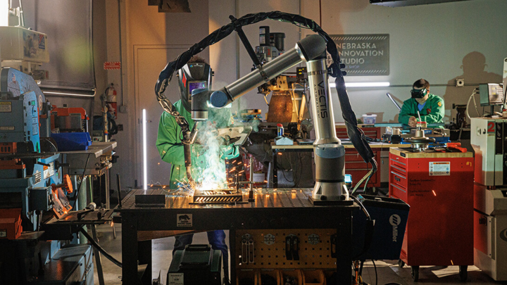 A Nebraska Innovation Studio member watches the welding robot while checking the programming on a tablet.