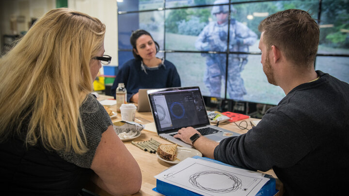 Veteran Melissa Ewing (left), Kendra Clapp Olguin, Has Heart communications director, and Husker alum Jordan Lambrecht work on the artwork representing Ewing's life story. Behind them, on screens at Nebraska Innovation Studio, is a photograph from Ewing's tour in Afghanistan. 